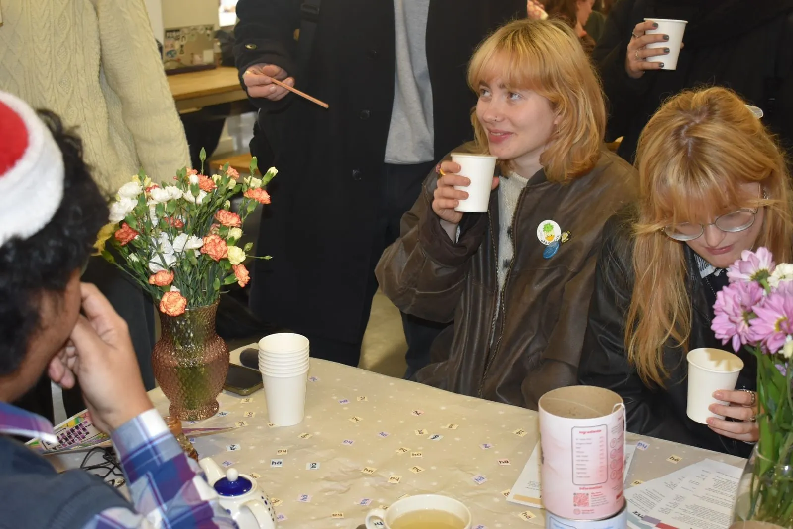 Students at a table at the launch, drinking Habu tea from paper cups beside a vase of flowers and a tin.