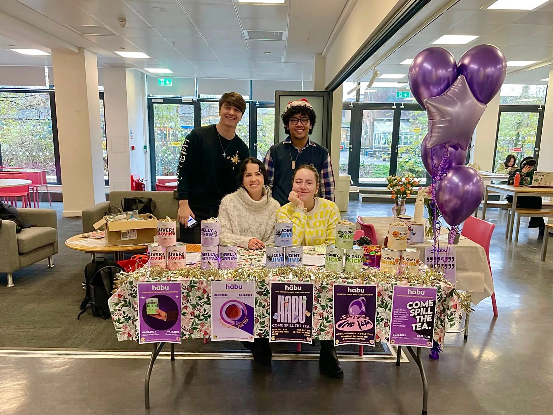 The Habu stall at the campus launch, a table of tins and posters under purple balloons, with the team behind it.