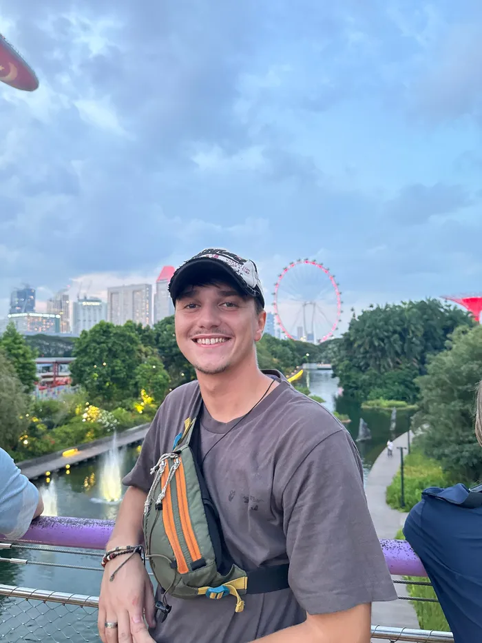 Denis smiling on a bridge with a city skyline and a big wheel behind him.