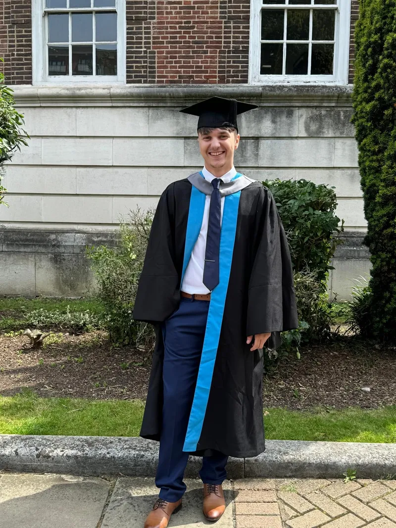 Denis in his graduation gown and cap outside a brick university building.