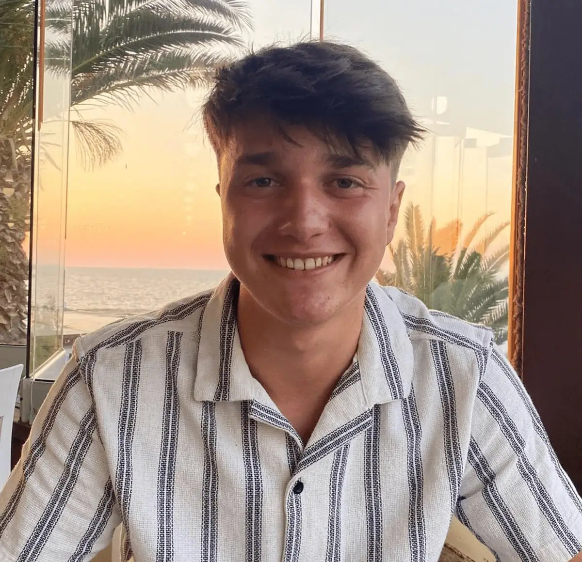 Denis smiling at a table by the sea at sunset, in a striped shirt.