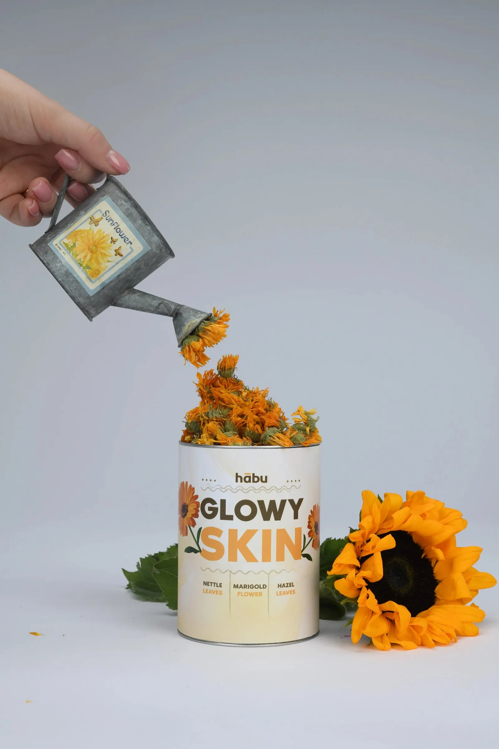 A hand pouring marigold petals from a small watering can into a Glowy Skin tin, with a sunflower beside it.